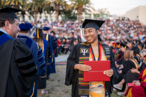 A graduate in cap and gown smiles as she holds her diploma cover