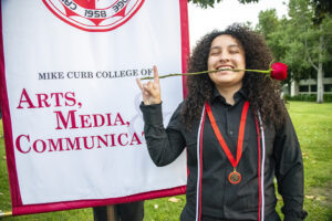 A graduate grins and holds a red rose between her teeth, and stands in front of a banner that reads: Mike Curb College of Arts, Media, and Communication.