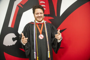 AS President Shayan Moshtael, in black robe, hood and honors cords, stands in front of a Matador mural in the University Library.