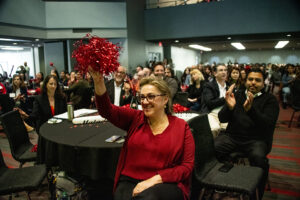 Audience sits in chairs and hold red pom poms in the air.