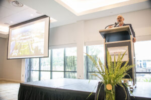 Colin Donahue stands on stage at podium with a screen to his left.