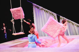 Characters in the play "Lysistrata" toss away fluffy pink pillows decorating seats in a TV studio set.