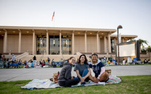 Three young women sit on a blanket on the University Library Lawn, they are smiling at the camera. People behind them are on the grass in front of a movie screen