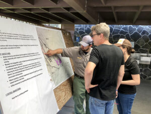 Mike Theune stands with two people in front of a large poster and a large map.