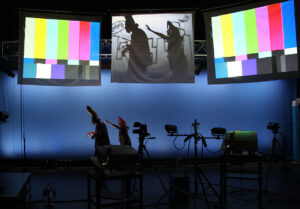 An actor chases another actor on stage while their images are also broadcast above them.