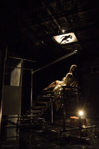 An actor posing as a body is silhouetted high in the lights above an actor onstage.