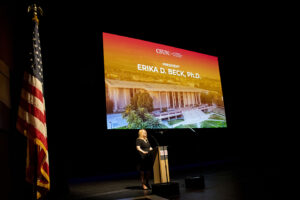 President Beck stands at podium on dark stage with a large screen behind her that displays her name,.