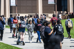 Students walk on the sidewalk near the University Library, which is in the background. Two students ride scooters amidst the pedestrians.