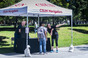 Three people stand under a "CSUN Navigators" pop up tent. The woman in the foreground speaks to a student and points, giving directions to the student.