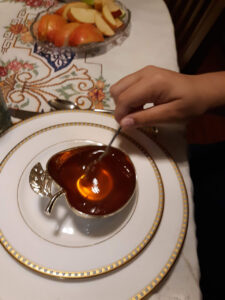 A hand stirs honey in a silver, apple-shaped bowl. The bowl sits on white china plates, with a plate of sliced apples sitting nearby.