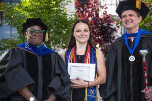 Tania Parker stands smiling and holding a certificate, with faculty members on either side of her.