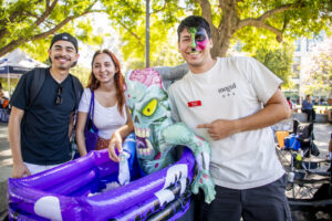 Two students at the "Zombie Apocalypse Scavenger Hunt" take a picture with an inflatable zombie in a coffin and an event worker with neon zombie makeup.