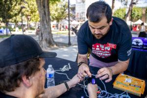 Daniel Ramos leans over a table, holding a cord to show a seated person how to make a bracelet.