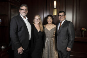 Four people stand together, arm and arm and smile at the camera