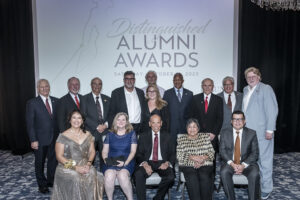 Group stands and is seated in front of a screen that says "Distinguished Alumni Awards."