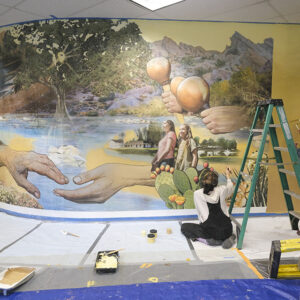 Artist Audrey Higa sits on the ground in front of the mural painting with small brush while Muralist Lindsay Carron works on the side.