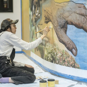 Artist with brush sits on the ground in front of mural, painting