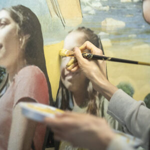 Close-up view of hands holding paintbrush and paint palette painting fine detail of people on mural.