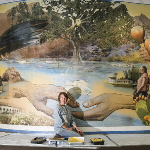 Lindsay Carron sits on the ground in front of her mural and looks at the camera.