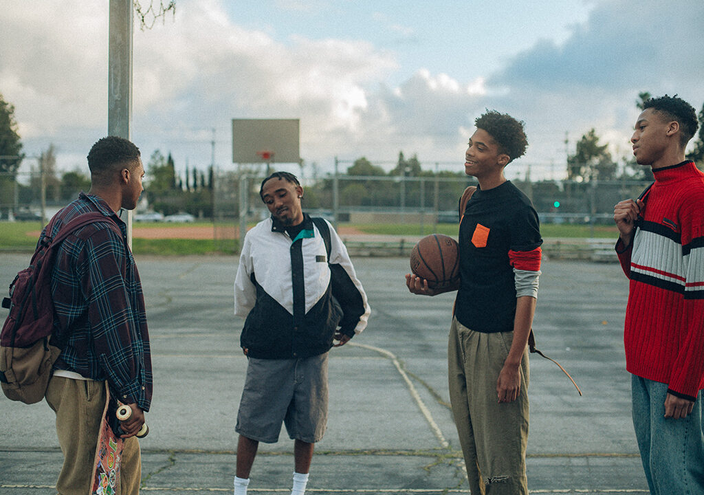 Four young men standing on an outdoor basketball court. One is holding a basketball