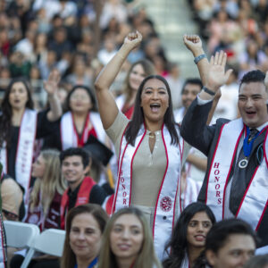 Graduates wearing white honors sashes cheer in the crowd.