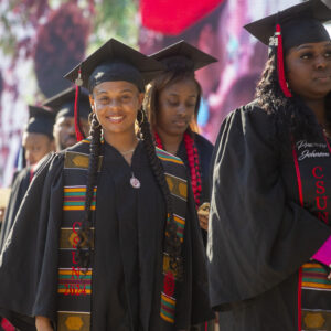 Students dressed in caps and gowns with colorful sashes stand in line. One graduate smiles at the camera.
