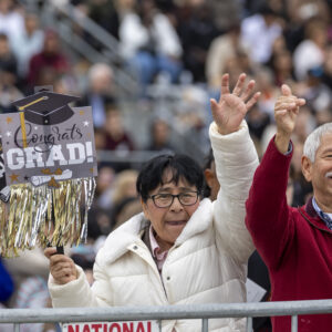 A couple waves to someone. One is holding "congrats grad" signs and the other holds up a cell phone to take a photo.