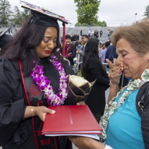 A graduate in cap and gown tearfully shows a woman her red diploma holder.
