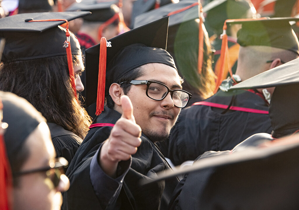 Student in cap and gown at a commencement ceremony looking at camera and give a thumbs up.