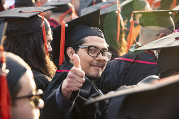 Student in cap and gown at a commencement ceremony looking at camera and give a thumbs up.