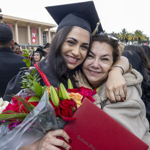 A joyful graduate hugs her mom and holds her red diploma cover and bouquet of flowers, after the commencement ceremony for CSUN's David Nazarian College of Business and Economics.