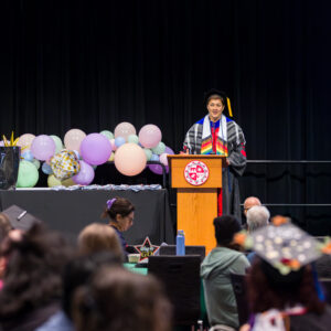 Sheena Malhotra stands in academic regalia at a podium on stage.
