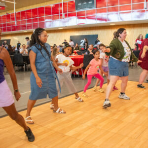 Group of people dancing on the dance floor at event with people sitting at tables behind them, watching them dance.
