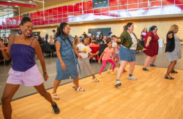 Group of people dancing on the dance floor at event with people sitting at tables behind them, watching them dance.