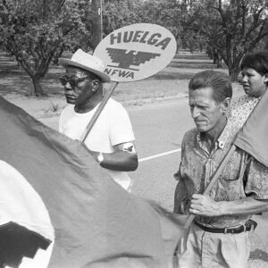 Two men, carrying signs and a UFW flag walking behind a UFW flag.