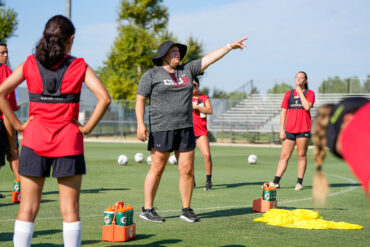 Gina Brewer stands in the center of a circle of the CSUN Women's Soccer team at a scrimmage game, pointing in front of her.