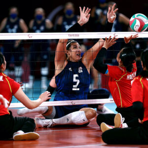 Katie Holloway Bridge, sitting, reaches up over a net to block a volleyball, in a sitting volleyball Paralympics competition.