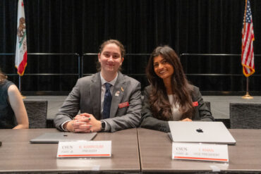 Associated Students President Katie Karroum and Vice President Iman Khan sit at a table behind signs that read their names and roles.