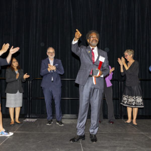 Dr. William Watkins stands on stage in the center of a six person half circle, pointing to the audience and smiling as he receives his award for 50 years of service.