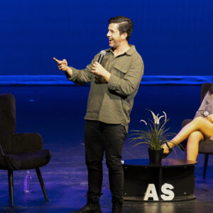 Actor Josh Peck stands on stage, smiling, pointing to the right.