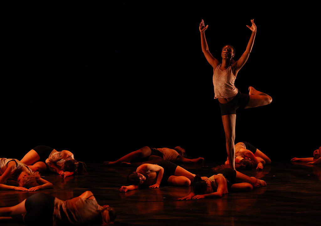 Image of one dancer standing with arms upraised while others lay before him.