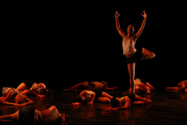 Image of one dancer standing with arms upraised while others lay before him.