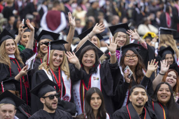 Students in caps and gowns smile and wave at the camera at Commencement, 2024.
