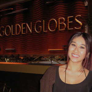CSUN film student, Nikolet Ocampo, smiles during her first Golden Globes rehearsal on Jan. 3, standing in front of the iconic "Golden Globes" stage signage. She is casually dressed in a black top, maroon pants, and a brown cardigan, wearing a production badge.