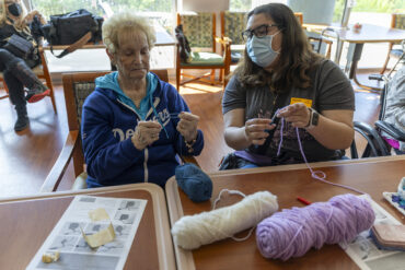 Two people sit side by side with crochet hooks and yarn.
