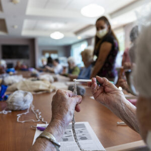 Hands hold a crochet hook and yarn while others look on in the background.