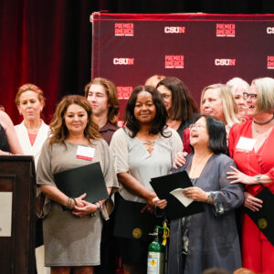 Women and family members from the 1987 softball team stand on stage, holding their certificates, as a teammate speaks at the podium.