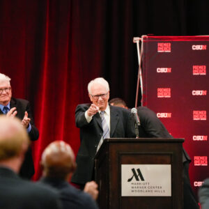 Don Strametz stands at the podium on stage, next to Master of Ceremonies Bill Courtland who is applauding him, pointing to the camera.