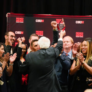 Don Strametz faces the back of the stage as he holds up his award in triumph and track and field alumni surround him applauding.