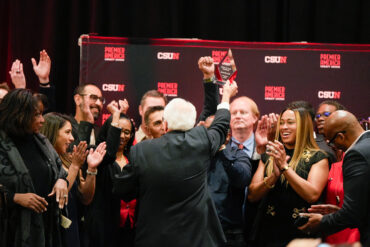 Don Strametz faces the back of the stage as he holds up his award in triumph and track and field alumni surround him applauding.